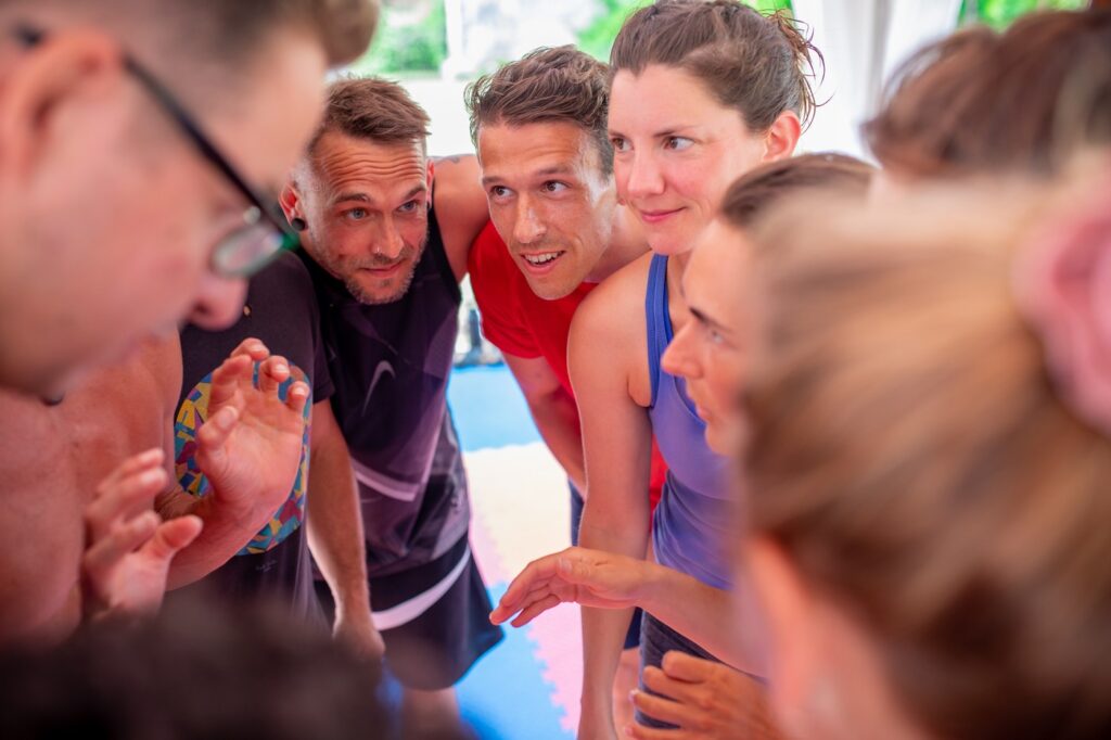 Group of people standing close, holding their shoulders while discussing an AcroYoga exercise.