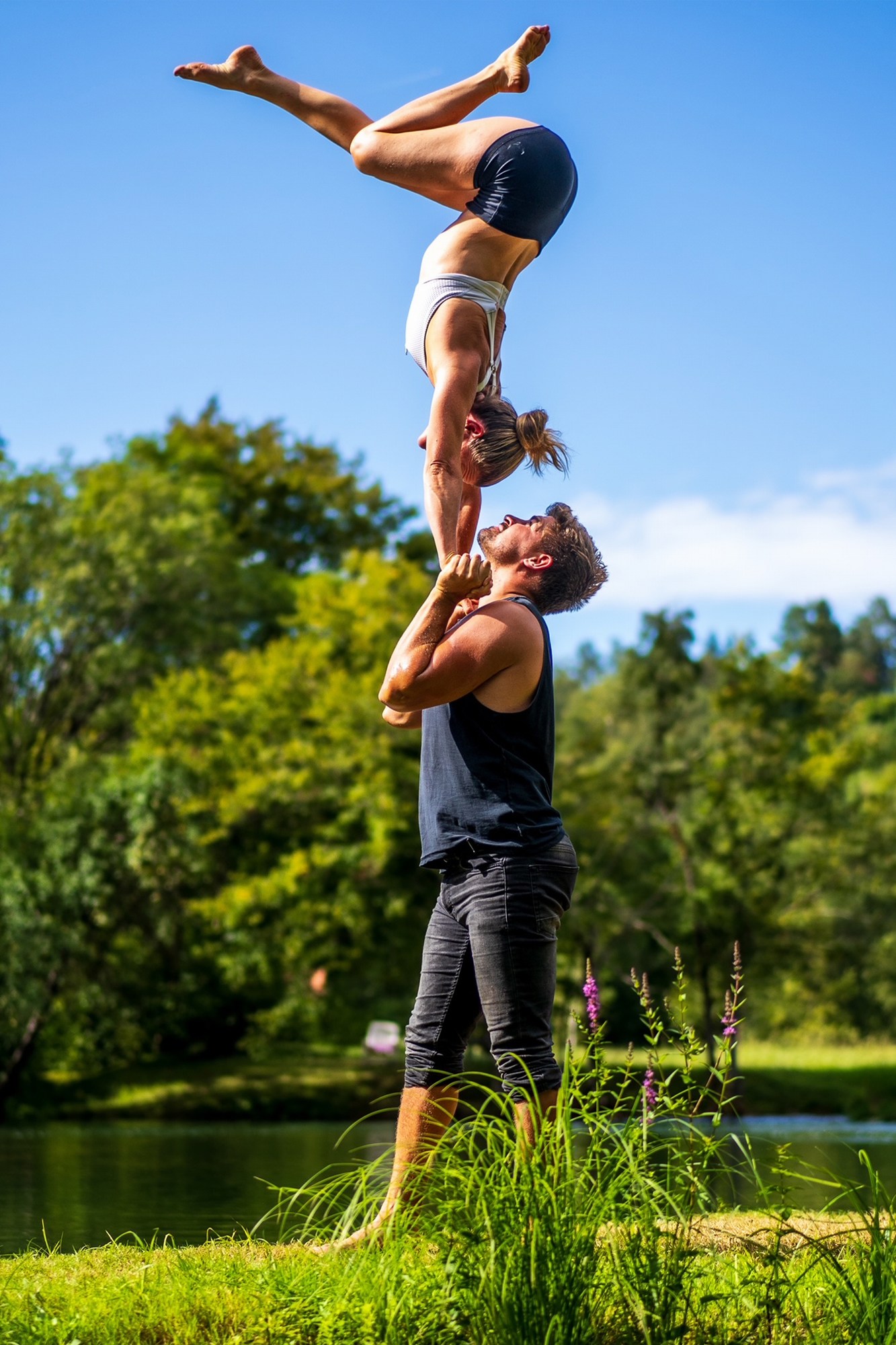 Girl being upside down in a handstand, balancing on a guys hand overlooking a green landscape and lake during a hot summer day.