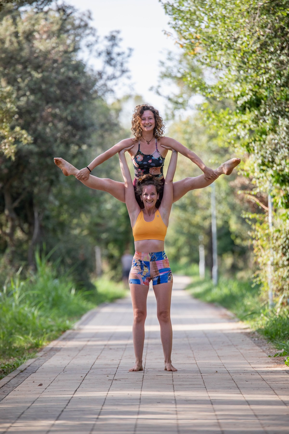 One girl basing another girl in a partner acrobatic pose high up in the air on a path in the woods.