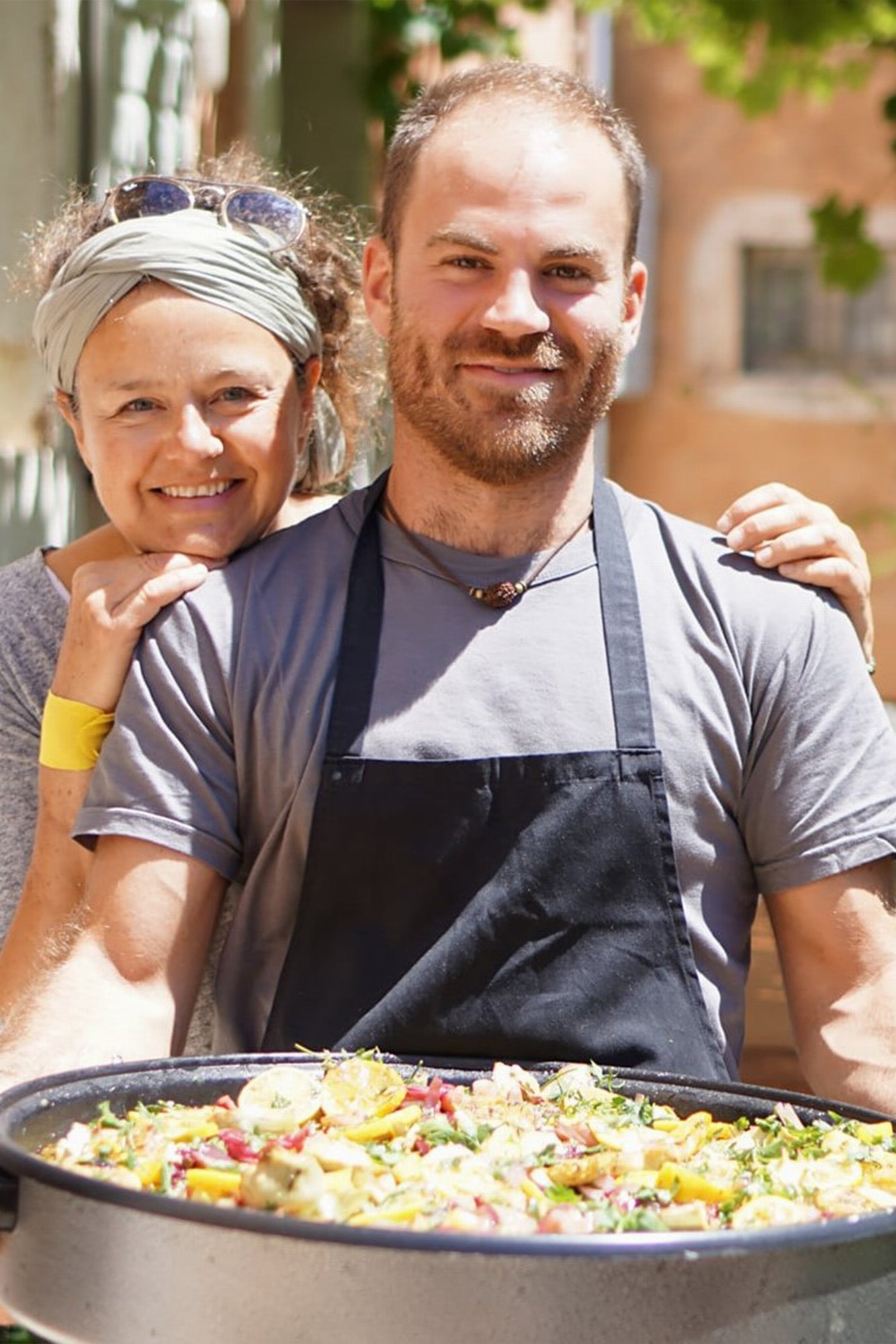 Image of two people smiling into the camera holding a big pan of fried rice with vegetables.