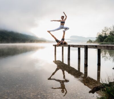 Girl in an AcroYoga pose on a wooden platform overlooking a morning scenery at the lake.