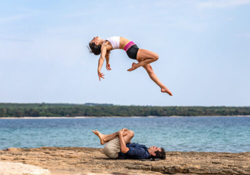 Couple doing AcroYoga, girl flying high in the air flying above the sea while her partner lays on the ground underneath waiting to catch her.