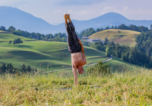 Guy being in a one arm handstand on grass with the background of green and lush hills in Slovenia.