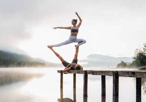 Girl in an AcroYoga pose on a wooden platform overlooking a morning scenery at the lake.