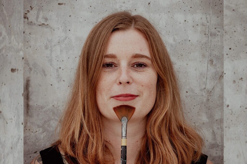 A woman with brown ginger hair smiling into the camera, standing infront of a cemet wall.